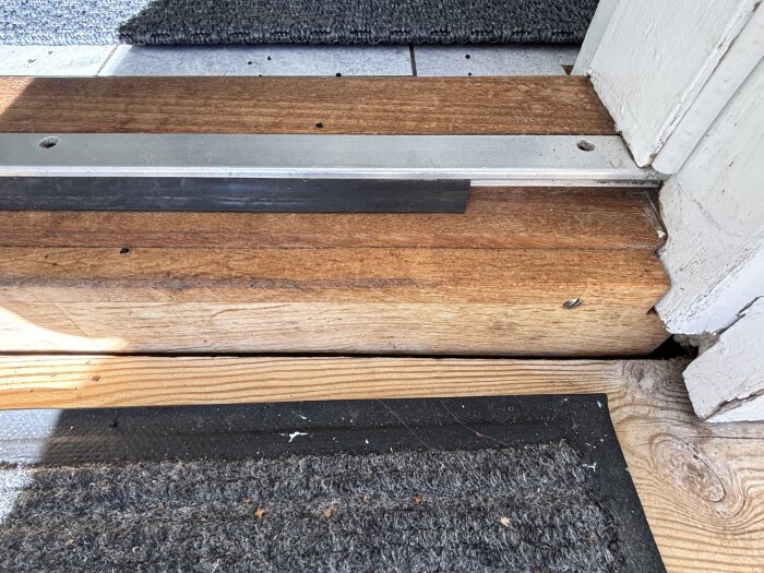 Sliten tröskeltätningslist av gummi på en äldre tröskel, delvis monterad längs en metallremsa, omgiven av trädetaljer och en matta.