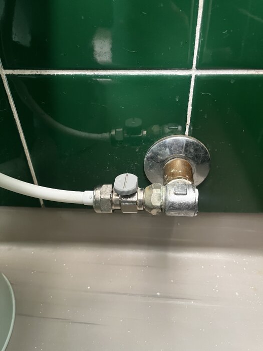 Vattenkoppling med vit slang och ventil, monterad mot en grön kaklad vägg, del av VVS-problem relaterat till flottör och vattenpåfyllning.