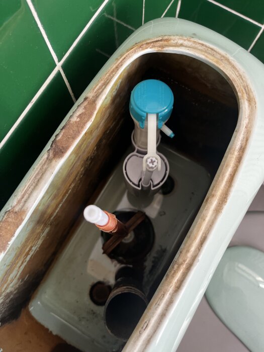 Närbild av en toalettcistern med flottör utan vatten, visar den blå flottörarmen och kopparrör inuti en vit porslinstank. Grönt kakel i bakgrunden.