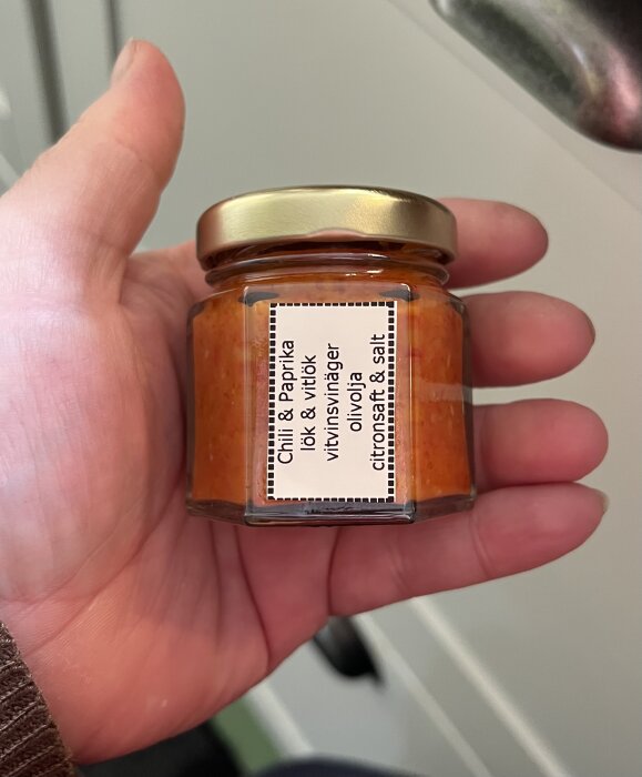 Liten glasburk med guldfärgat lock, innehållande hemmagjord tomat och paprikamarmelad med chili. En hand håller burken.