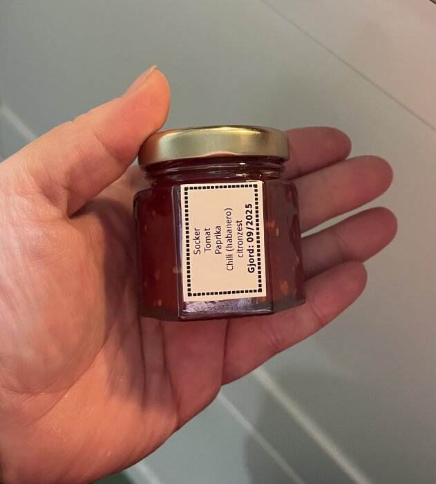 Hand som håller en glasburk med etikett som visar ingredienser: socker, tomat, paprika, chili och citronzest, datum 09/2023.