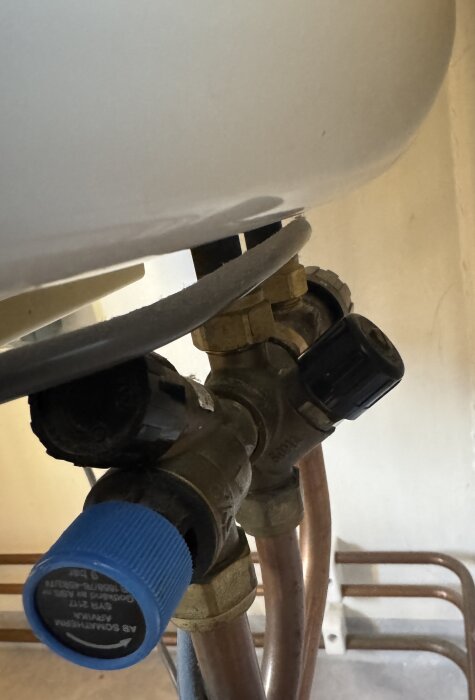 Närbild av rörkopplingar under en varmvattenberedare med ventiler, en med blått handtag och en utan, med kopparrör anslutna.