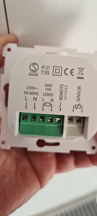 Termostatens baksida med anslutningar och symboler för kablar, inklusive last, fjärranslutning och sensor. Hållen i handen, vit bakgrund.