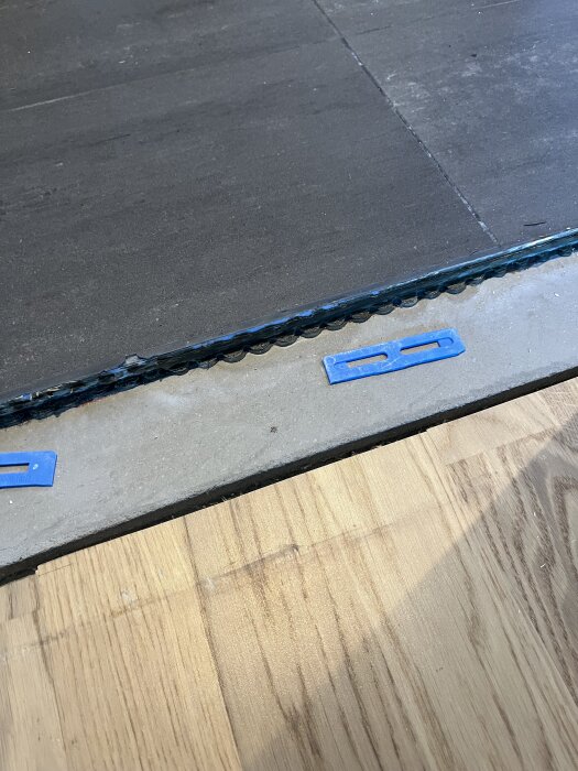 Borttagen tröskel med exponerad betongyta och övergång mellan trägolv och mörkare yta, med blå distansbrickor synliga.