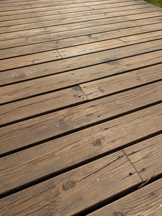 Närbild av ett trädäck med slitage och tecken på tidigare behandling, möjligen med rinnmärken, beläget under ett altantak.