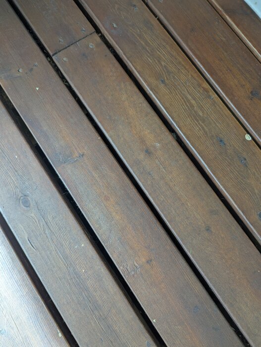 Bruna träplankor på altandäck med synliga skruvar och några små märken, troligen med tecken på tidigare behandling.