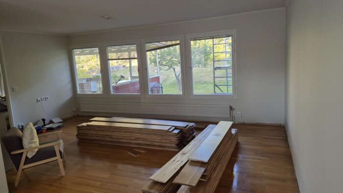 Rum med stora glasfönster, gammal radiator under fönstren och staplade träplankor på golvet för renovering.