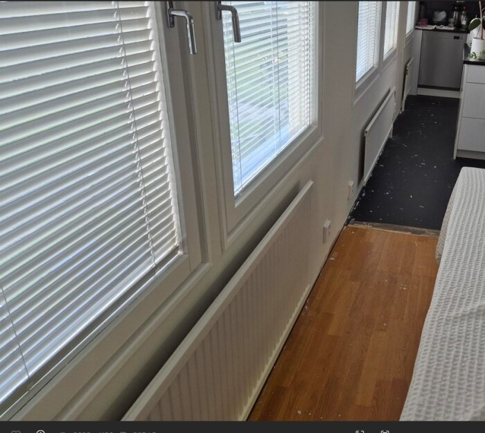 Radiator under tvåglasfönster i vardagsrum med parkettgolv, del av kök synligt i bakgrunden, fönster har persienner och utsikt mot trädgård.