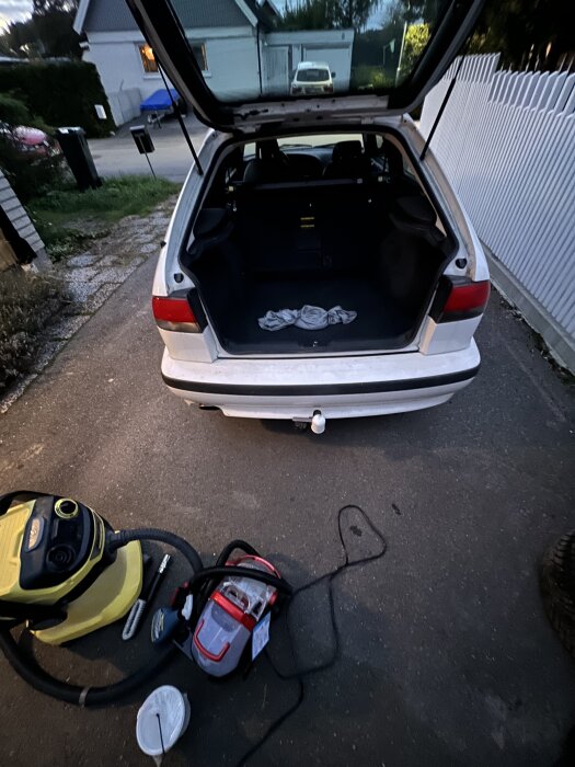 Öppen baklucka på en vit bil med en dammsugare och rengöringsutrustning på marken, för sanering av mögel i bilen.