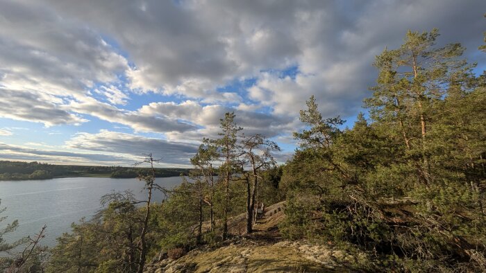 Vy över en sjö och barrskog från ett berg under en himmel med dramatiska molnformationer och blåa partier.