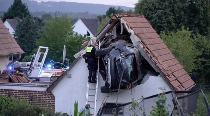 En bil har kört in i ett hus och fastnat i taket. En räddningsarbetare på en stege undersöker bilen.