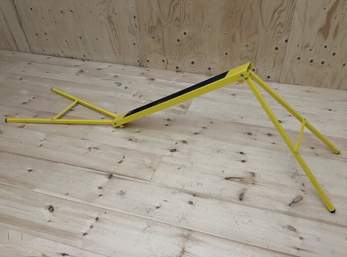 Gul justerbar arbetsbock i metall med svart ovansida, placerad på ett ljust trägolv, mot en träpanelvägg i bakgrunden.