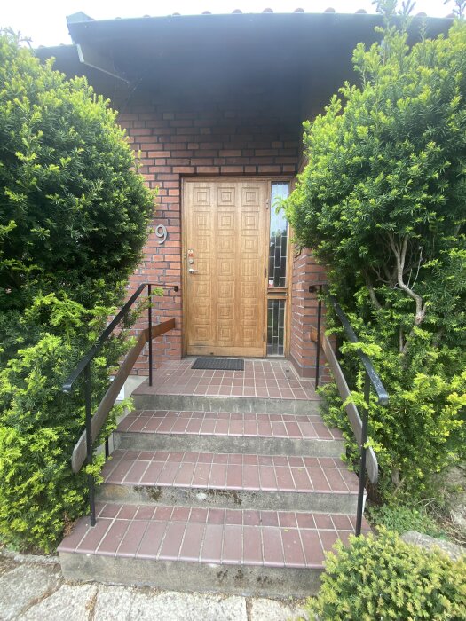 Entré till 60-talshus med trädörr, tegelväggar, trappsteg i rödbrun sten, omgiven av frodig grönska.