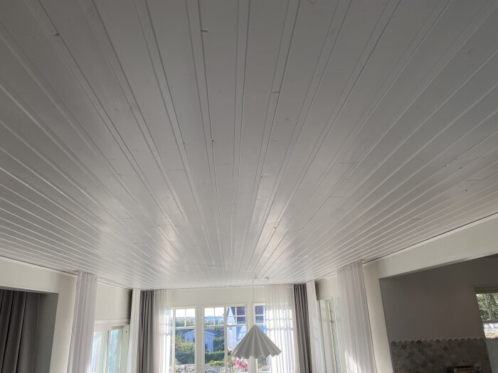 Vardagsrum med vitt paneltak, stora fönster med gardiner och en lampa. Diskussion om att byta tak för en jämn yta.