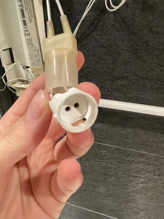 En hand håller en kopplingskontakt för lysrör med ledningar, mot en bakgrund av ett grått golv och en del av en armatur.