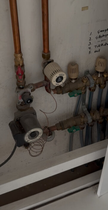 Rör och ventiler för vattenburen golvvärme med nummermärkta justeringsanordningar på en vägg i källarplan.