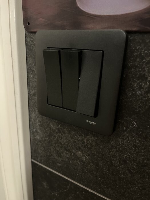 Svart Schneider Exxact strömbrytare med tre knappar monterad på en mörk kakelvägg.