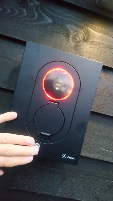 Zaptec laddbox visar felkod och röd ljusring; hand håller över en QR-kod på boxens framsida, monterad på mörkt trä.