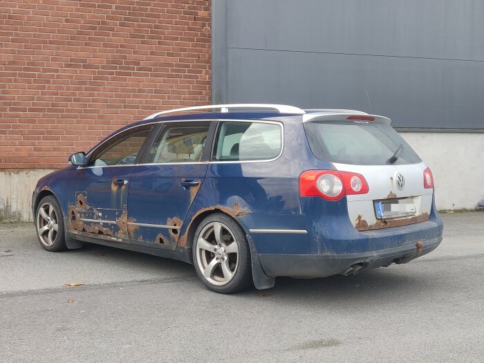 En blå Volkswagen-kombi med rostskador står parkerad utomhus framför en tegelvägg.