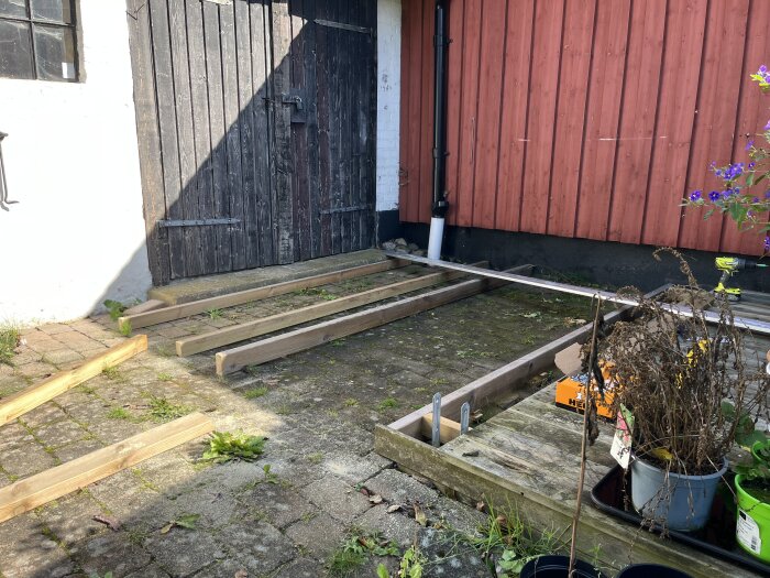 Byggprojekt vid uteplats med lagda plankor på ojämnt stenlagd mark nära en dörr på en röd trävägg, omgivna av byggmaterial.