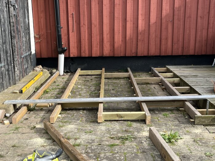 Byggprojekt av altangolv med träbalkar över ojämnt stenlagd yta, mot en rödvägg med stuprör, nära en befintlig veranda.