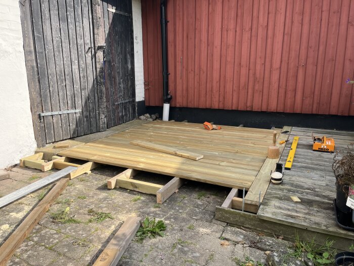 Nysnickrad uteplats med trallbrädor framför en röd trävägg. En vattenpass och verktyg syns ovanpå den halvfärdiga konstruktionen.