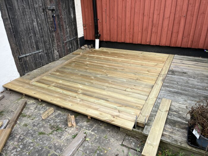 Träaltan under konstruktion, delvis färdig, med nyare brädor över gamla stenplattor bredvid en röd trävägg och en svart tappkran.