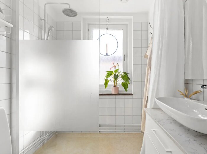 Modernt badrum med en dusch bakom en fast glasskärm, vitt kakel och ett handfat bredvid en växt i fönstret.