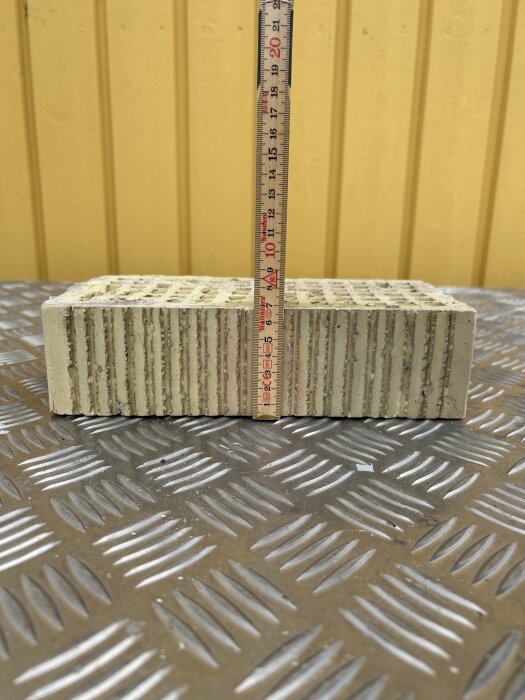 Tegelsten stående på metallgolv med en måttstock som visar höjden framför en gul vägg.