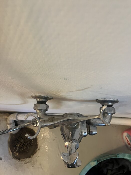 Väggmonterad kromblandare med anslutna rör syns ovanför ett avloppsbrunn på golvet.