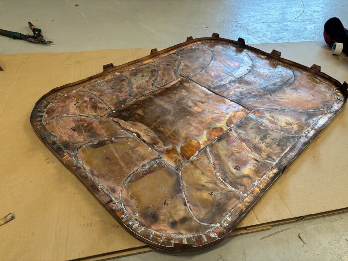 Kopparplåt med lödda bitar och vågformad yta, placerad på kartong, omgiven av verktyg och golv.