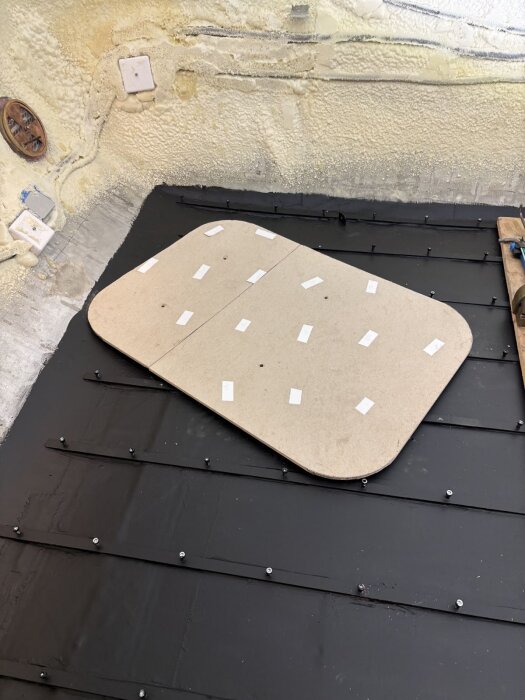 Arbetsyta med svart plåt och en monterad fiberskiva med tejp. Skivans kanter överlappar plåtens svarta yta. Vit isolering syns i bakgrunden.