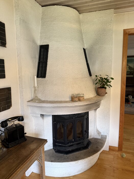 Vit braskamin med svart glaslucka och ventiler, omgiven av en hylla med växt och ljus, står i hörnet av ett rum med trägolv och träpaneler.