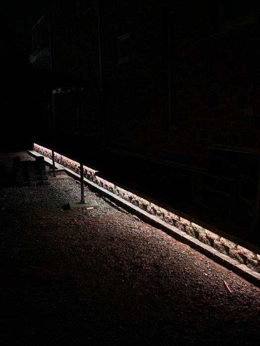 Mörk bild av en kantsten längs en byggnadsvägg, belyst nedifrån med rödaktigt ljus, omgiven av grus på marken.