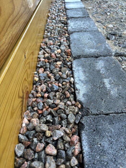 Kantsten med dekorativ småsten längs en trävägg, del av ett byggprojekt med fokus på trädgårdsdesign och utrymmen.