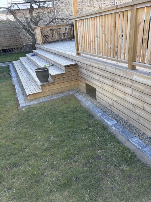 Träaltan med trappa, kantsten och dekorsten ovan gräsmatta, med räcke omkring och krukväxt på trappan.