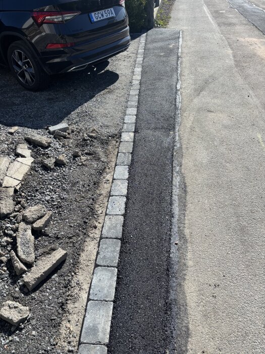 Bilen står parkerad vid en nyanlagd asfaltkant med kantstenar längs trottoaren. Grus och stenar syns vid sidan av vägen.