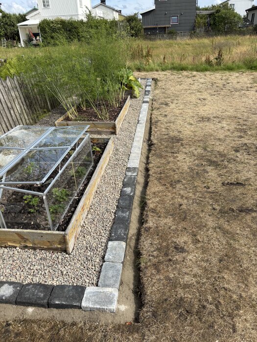 Odlingsyta med kantsten och grus, omgiven av gräsmatta. På odlingsytan finns även en träbädd med glastäckta växter.