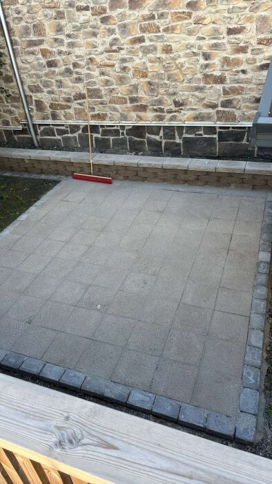 Markyta med nylagd marksten och kantsten intill en stenmur, med en röd sopkvast uppställd mot muren.