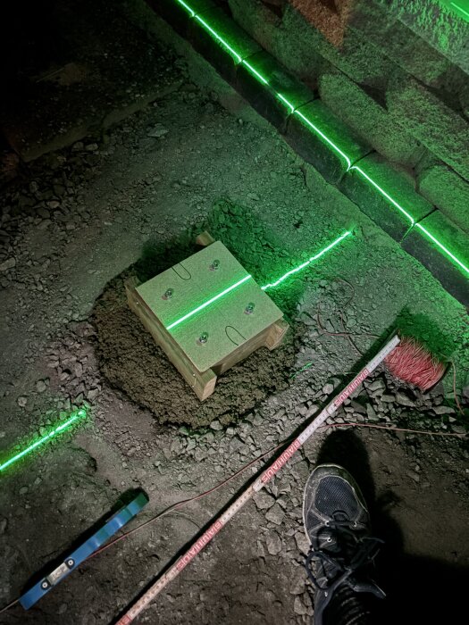 Personliga skyddsskor framför kantsten med grön laserlinje och träform för gjutning i grusig markmiljö.