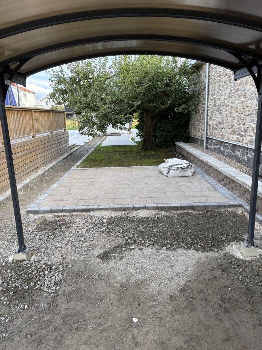 Carport med marksten och kantsten, träplank och stenmursyta i trädgård, grön växtlighet bakom och pågående markarbete i förgrunden.