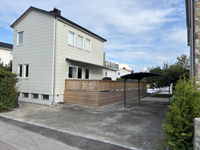 En tvåvåningsvilla med carport, kantsten och trädgård. Renoveringsprojekt med ny altan och marksten. Solig dag med blå himmel.