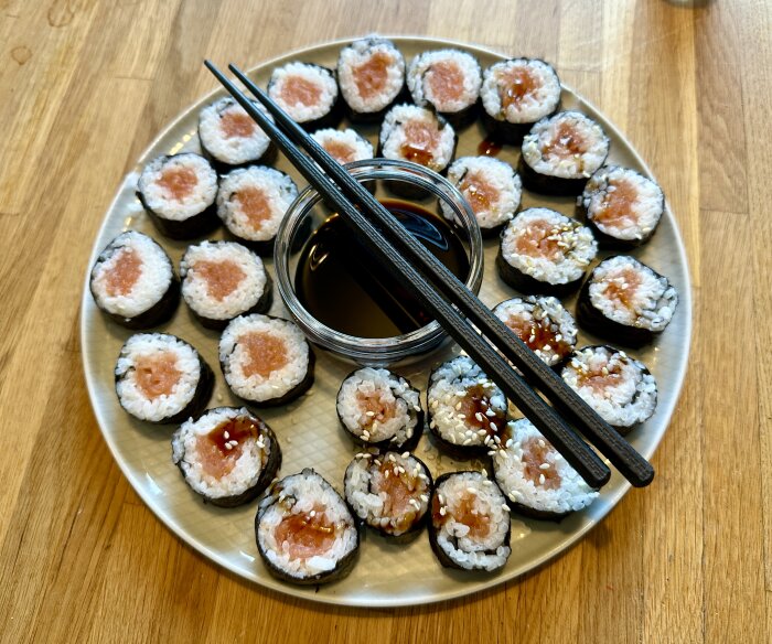 En tallrik med sushibitar, troligen makirullar, fyllda med ris och lax. I mitten står en skål med soja och svarta ätpinnar ligger ovanpå.