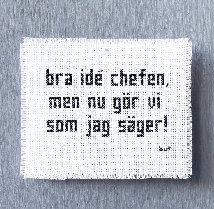 Broderi med texten "bra idé chefen, men nu gör vi som jag säger!" monterad på en grå vägg.