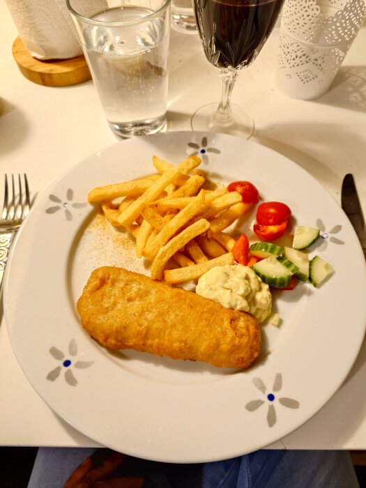 Tallrik med fish and chips, pommes frites, tartarsås och sallad. Bredvid står ett glas vatten och ett glas röd dryck.