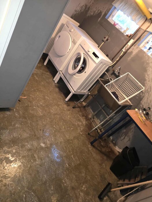 Tvättstuga med en tvättmaskin och torktumlare, fuktig golvyta, diskho, och ett fönster med litet ljusinsläpp.