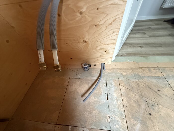 Orenoverat badrum med obehandlad trägolv och vägg, dragna rör i hörnet.