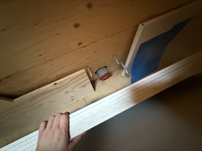 Ovanvåningens badrum under renovering, visar trävänster, rör och blå skyddspapp. Hand håller vit byggskiva.