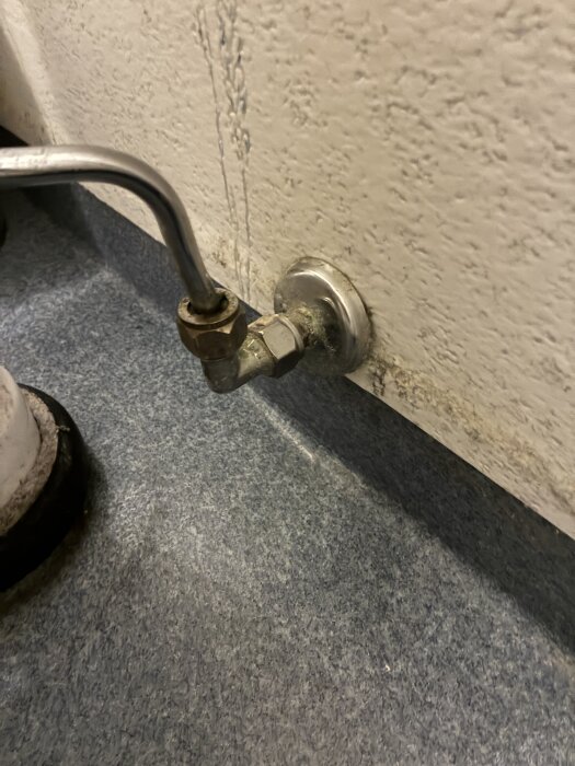 Röranslutning på väggen med metallbeslag och gängad koppling, monterad mot texturerad yta och liggande på ett golv.