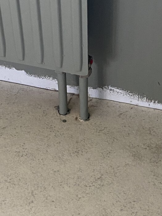 Grå radiator med två rör som leder från botten av elementet ner till golvet, cirka 3-4 cm från vägg och 2 cm mellanrum mellan rören.